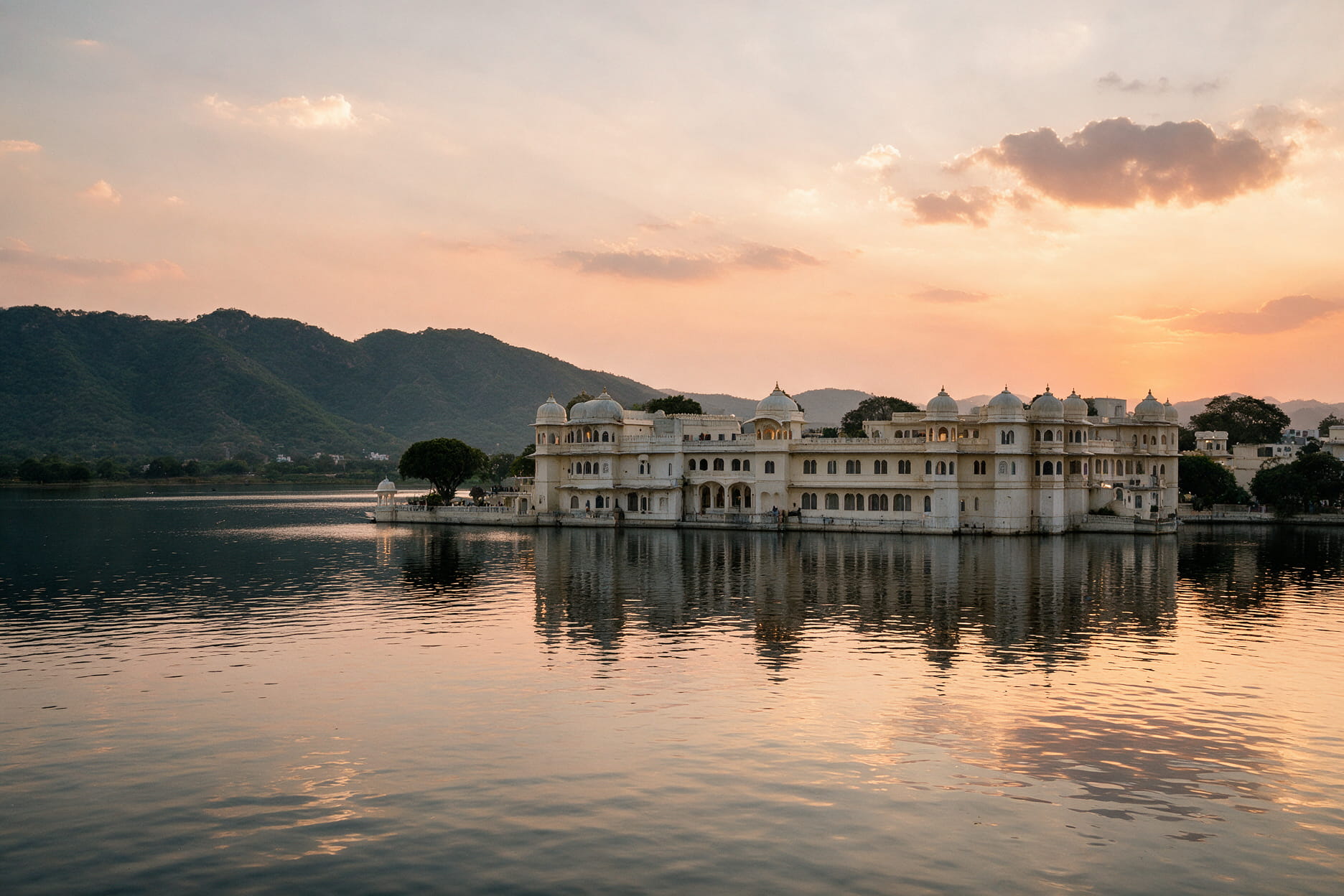 Lake Pichola at sunset with the City Palace reflecting on the water and green Aravalli hills in the background.