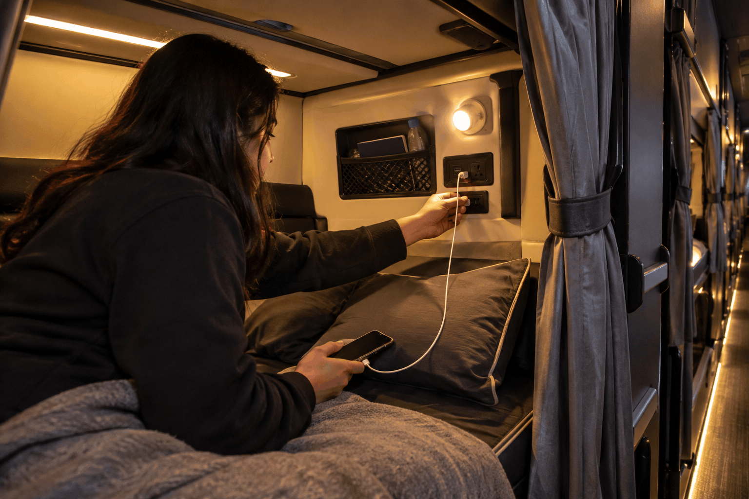 Passenger plugging a phone cable into a charging port beside a sleeper berth on an overnight bus